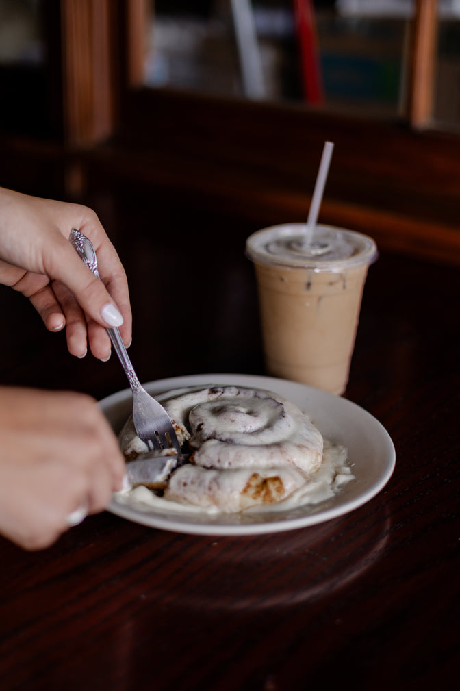 Person cutting into a cinnamon roll with a fork on a plate, next to an iced coffee drink.