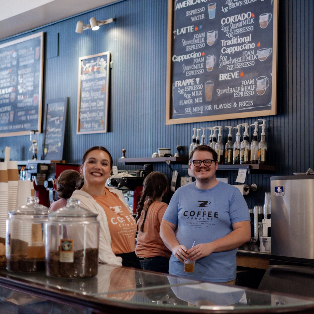Two people standing behind a counter in a coffee shop with coffee-making equipment and signs on the wall.