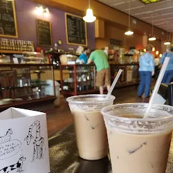 Two iced beverages in plastic cups with straws on a table in a coffee shop.